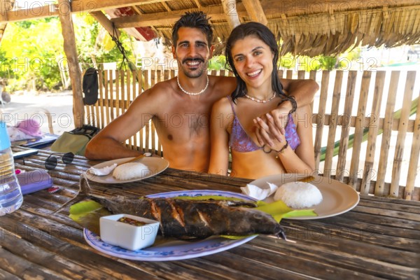 Tourists embracing local cuisine during their vacation in the philippines, savoring a grilled fish meal with rice in a beach hut