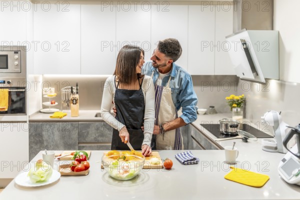 Young couple wearing aprons preparing a vegetable salad together in their modern kitchen, embracing and smiling