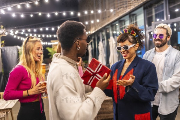 Multi-ethic friends giving a gift box to a woman celebrating birthday in the terrace of a modern bar at night