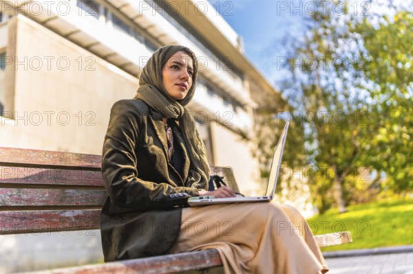 Side low angle view of a muslim businesswoman with headscarf working on remote sitting on park bench