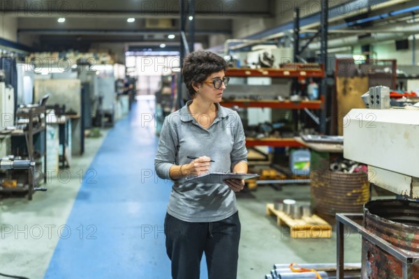 Female caucasian manager of a metallurgic cnc factory doing inventory holding a clipboard walking along an aisle