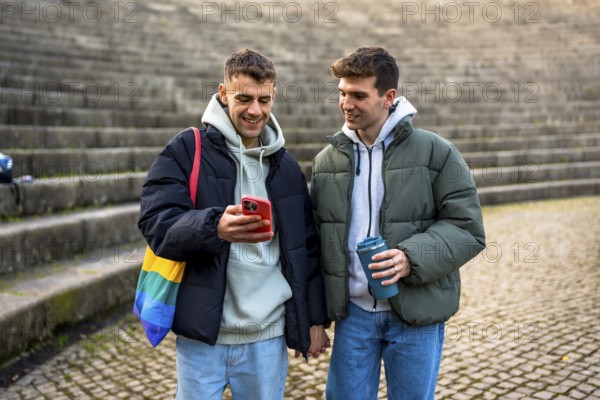 Happy gay couple holding hands, looking at smartphone and enjoying a reusable cup of coffee outdoors