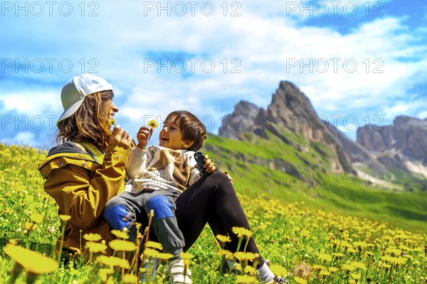 Mother and son playing with dandelions in a field of wildflowers with the seceda mountains in the background, enjoying summer in the italian dolomites
