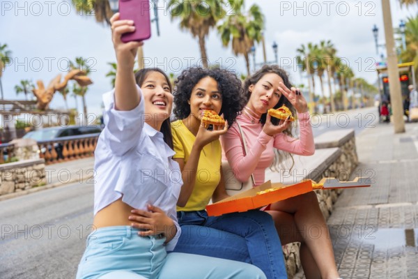 Multi-ethnic female friends taking a selfie while sharing pizza sitting in the street