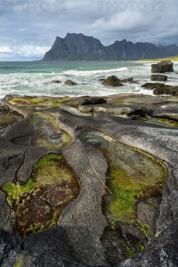 Seascape on the beach at Uttakleiv (Utakleiv), in the foreground rocks and rocky outcrops filled with water, including the so-called Eye of Uttakleiv, a round stone in the water. Mount Hogskolmen in the background. Sun and clouds. Early summer. Uttakleiv, Vestvagoya, Lofoten, Norway, Europe