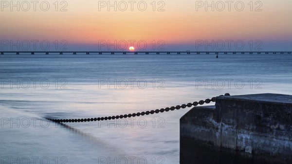 A serene sunset with the sun touching the horizon, beside a calm ocean and a pier extending into the distance, Lisbon