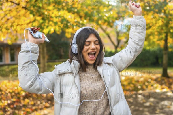 Happy vital caucasian young woman dancing listening to music in a park with fallen leaves in a sunny day of autumn