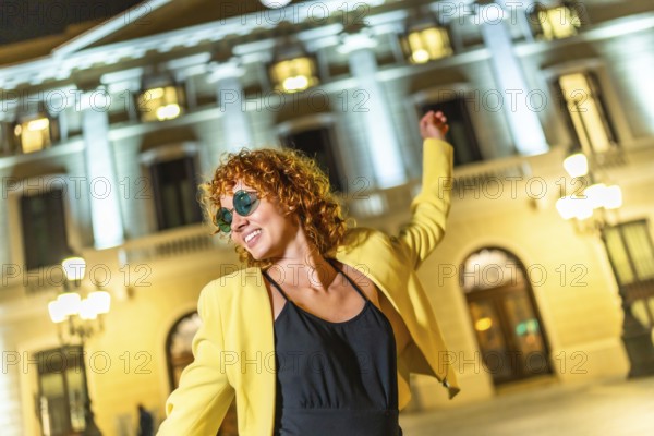 Happy woman in yellow jacket, black summer dress and sunglasses dancing alone in the city at night