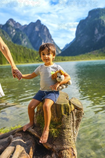 Young boy holding a plush toy and his mother's hand while sitting on a tree stump by lake dobbiaco, surrounded by the dolomites