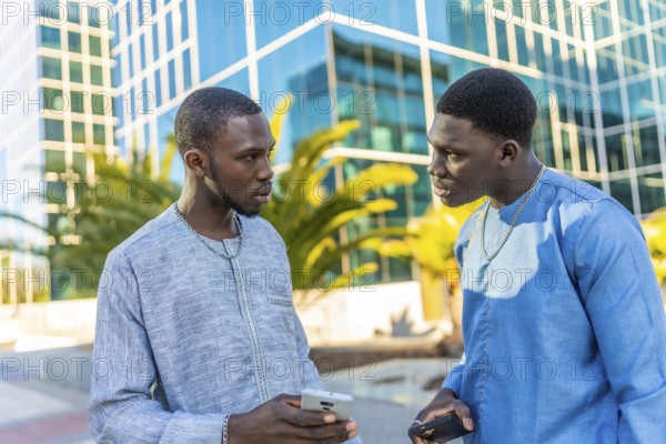 Two senegalese businessmen wearing traditional dashiki attire are discussing business while holding smartphones in front of modern office buildings