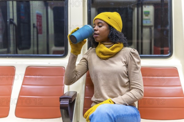Young woman wearing winter clothes is sitting on a subway train, enjoying a warm beverage from her reusable cup