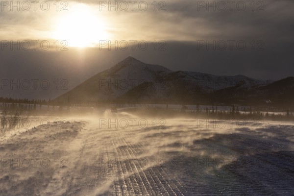 Snow-covered gravel road in the wind in front of mountains, backlight, ice, Dalton Highway, Alaska, USA, North America