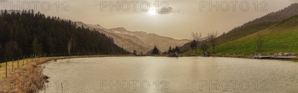 A tranquil lake against a mountain backdrop at sunset with trees and a gentle sky, the small Bichlersee lake