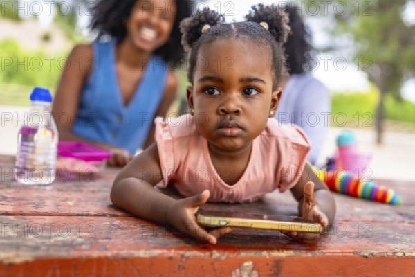 Close-up of a cute African baby girl using mobile phone in the park while her mother and granny talking on the background