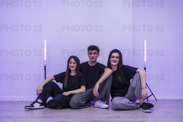 Three young dancers are sitting together on the floor of their dance studio, taking a break and enjoying the purple ambient lighting