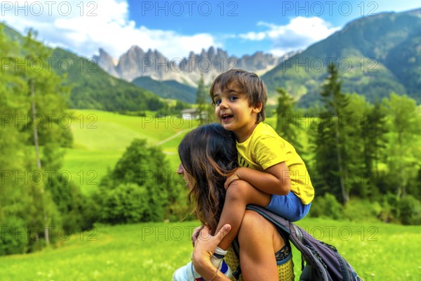 Mother carrying her son on her shoulders while enjoying the view of the odle mountains and the funes valley in the dolomites in summer