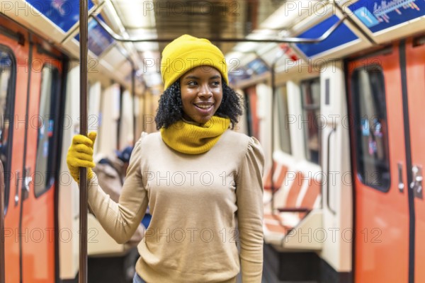 Young black woman wearing yellow wool cap and gloves standing inside subway train, holding handrail and smiling