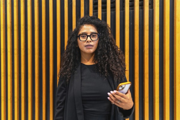 Portrait of a latin young businesswoman wearing eyeglasses and formal elegant black cltohes in a modern building holding a mobile phone and looking at camera
