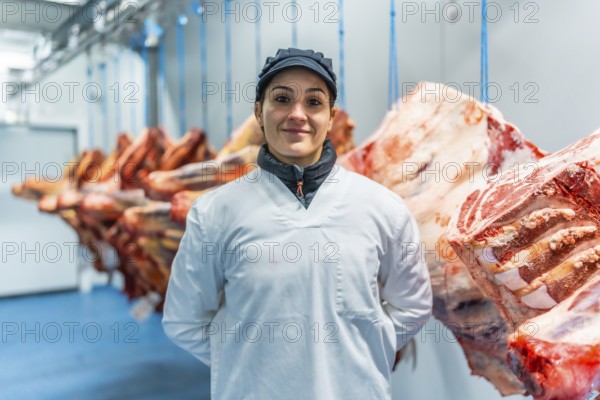 Frontal formal portrait of a happy caucasian adult woman butcher with laboratory coat and hygienic hat in a cold storage room of meat factory