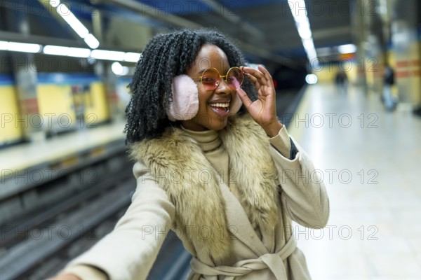 Smiling young woman wearing earmuffs and stylish glasses taking a selfie while waiting for the train in subway station