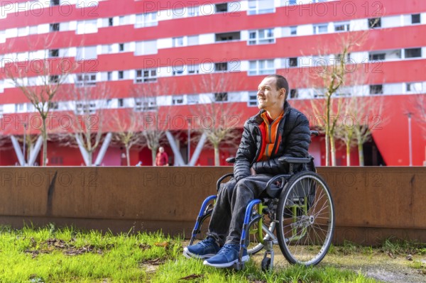 Young man sitting in wheelchair enjoying a sunny day in a modern urban park with a red building in the background