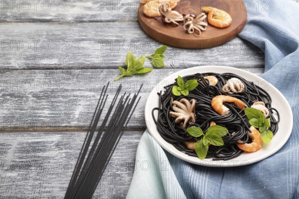 Black cuttlefish ink pasta with shrimps or prawns and small octopuses on gray wooden background and blue textile. Side view, copy space