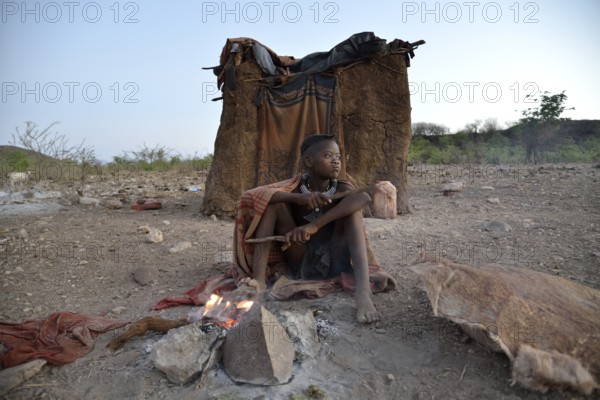Young shepherdess from the Himba people sitting at a campfire in front of her hut, Ombombo, Kaokoland, Kunene, Namibia, Africa