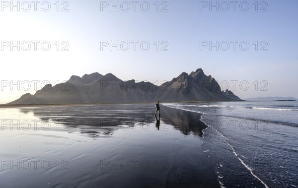 Tourist at black lava beach, sandy beach and sea, mountains Klifatindur reflected in the water, Eystrahorn and Kambhorn, headland Stokksnes, mountain range Klifatindur, Austurland, East Iceland, Iceland, Europe