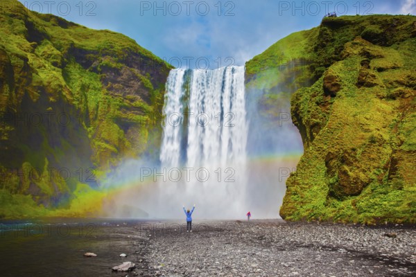Grand waterfall in Iceland - Skogafoss. Picturesque huge rainbow appears in the water mist. Middle-aged woman - tourist shocked beauty waterfall