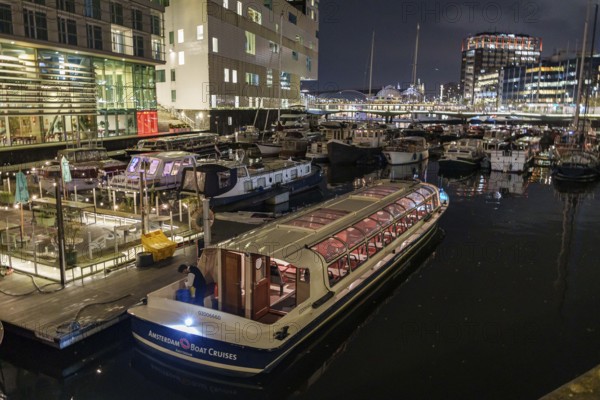 Illuminated boat in a harbour at night surrounded by modern buildings and reflections in the water, Amsterdam, Netherlands