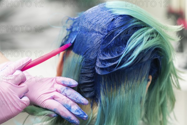 Side view of process of dyeing hair in unique color. Hairdresser in protective glove using pink brush while applying blue paint to female customer with emerald hair color