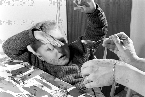 A boy is sick and won't take his medicine. His mother holds out a spoon and waits for him to gape and take it, but he tries to avoid the medicine by moving backwards and down. An everyday life event that today would look the same, the medicine sometimes does not taste so good. Sweden 1956. Conard ref 3157