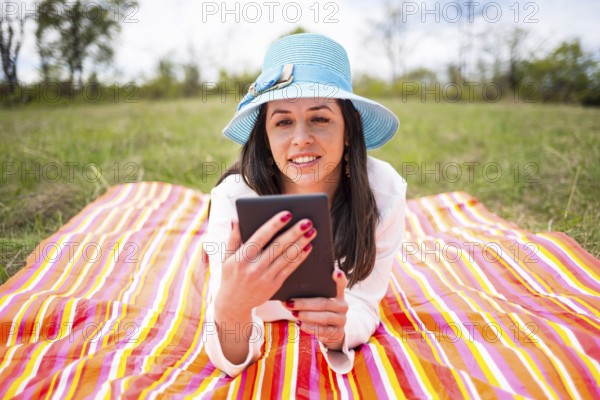 Attractive smiling young woman dressed casually uses e-book/tablet lying down on a colourful blanket in the park. Modern lifestyle using portable mobile devices everywhere people go. Reading e-mail