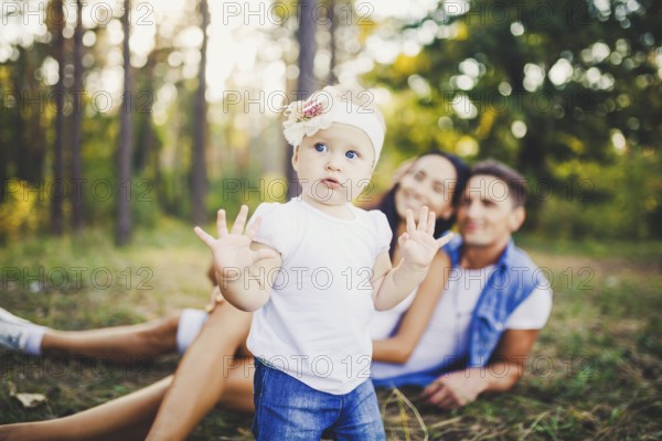 Little girl one year on the background of parents resting on graß learning to walk on nature in the park. The first steps of a child in nature outside the city in the forest