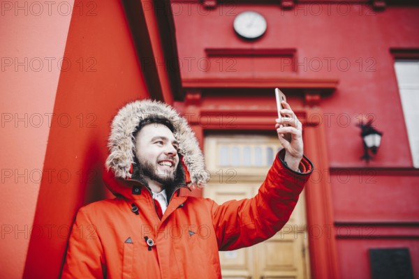 Handsome young male student with smile and beard stands on background red wall, facade of educational institution in red winter jacket with a hood with fur in winter. Uses a mobile phone, does selfie