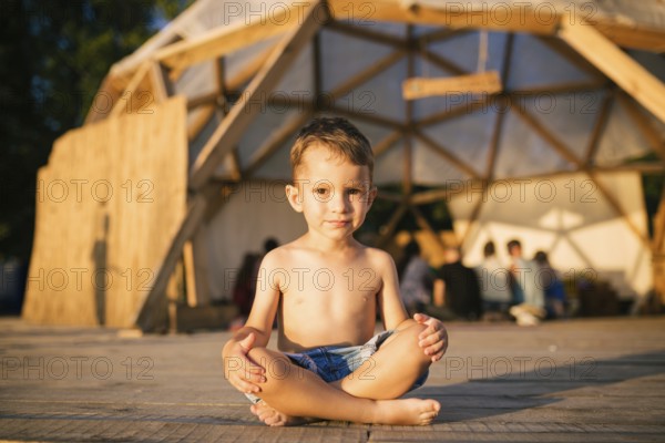 Theme is yoga and children. Caucasian Boy child sitting barefoot cross-legged in lotus position on wooden floor the background of gathering place of people. In summer sunset. Dressed in denim shorts