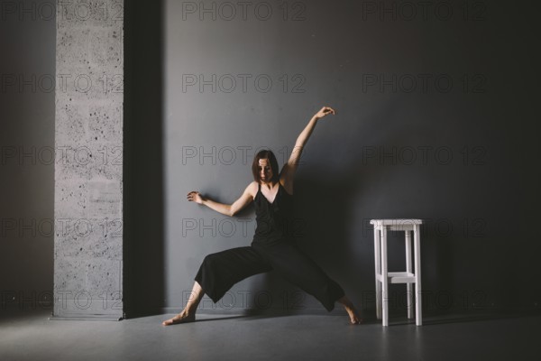 Social theme of female loneliness is pain suffering. Abastration of male domestic violence. Young beautiful Caucasian woman in black clothes, strange pose, white chair big dark room. Fear and despair