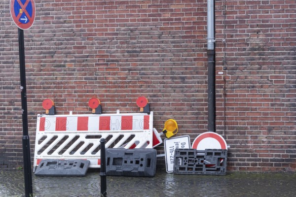 Red and white construction barrier, traffic signs, and bases stacked against a brick wall with no parking sign