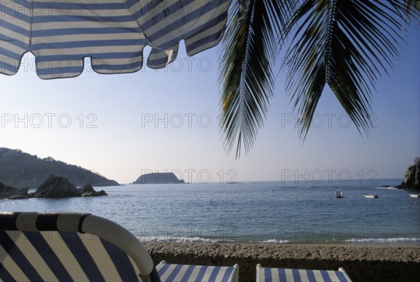 A tranquil beach scene with palm leaves, a striped umbrella, and a view of the ocean and distant island, in the coast of Mexico