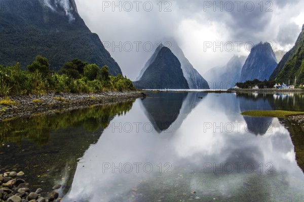 Mirror-smooth water of the Milford Sound fjord reflects mountains. Sunlight breaks through storm clouds. Land of goblins and hobbits. New Zealand. Concept of exotic, active and photographic tourism