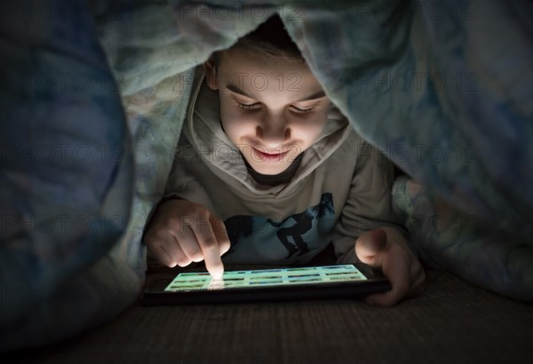 Child watching his tablet in the bed. Illuminated child face from device screen. Smiling Boy under the covers hold a tablet. Night time