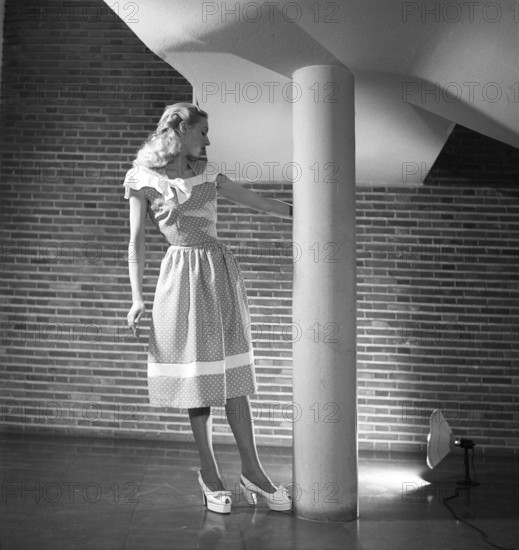 Fashion 1946, a young blonde female model dressed in the fashion of the year. Her sleeveless dress has a polka-dot pattern and is of a summery, lightweight type with a narrow waist and a length that reaches below the knees. A white band of fabric is repeated on both its top and bottom. Kristoffersson ref Y85-5