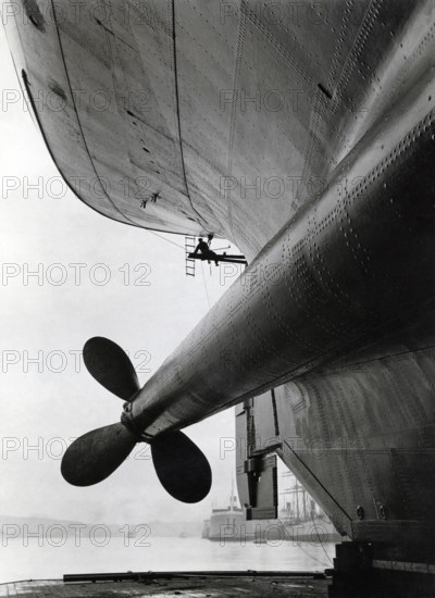 Dockyard in the 1930s. The ship M/S Gripsholm is undergoing maintance and the ship is risen in a dry dock. One of the stern propellers is visible and a worker is seen working on the hull. Theh ship was delivered 1925 to the swedish shipping company Svenska Amerika Linien and was at the time the most moderna ship on the Atlantic. It had a length of 168, 5 meter and 22, 7 meter wide. A veritble luxury palace in witch the most famous people travelled the seas. It was built in Newcaste England by Armstrong, Whitworth & Co