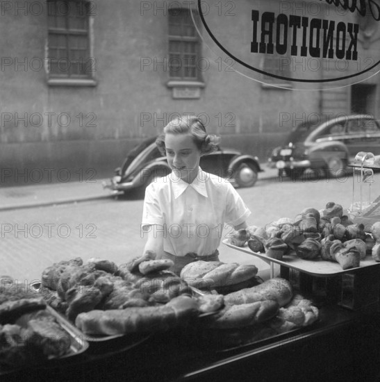 In the 1950s. A woman is pictured in the street and at the storefront of a bakery. She looks as if she is deciding what to buy and looks tempted by the good selection of sweet bread and buns. Sweden 1951 Kristoffersson ref BD91-5