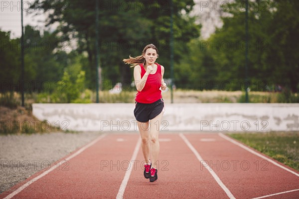Beautiful young athlete Caucasian woman with big breasts in red T-shirt and short shorts running jog, running in a stadium with a red rubber coating. Hairstyle of ponytail