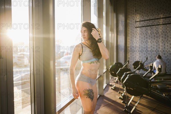 Young caucasian sporty woman with curly long hair poses in sportswear near a panorman window in a fitness club. Young female athlete model with beautiful body stands in the gym with a tattoo