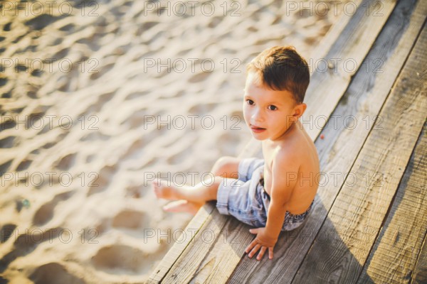 Little Caucasian boy child sitting on wooden pier sandy beach, summer time, sea holiday near water. The theme is the flow of time, a short life, the meaning and purpose of existence. Meaning of life