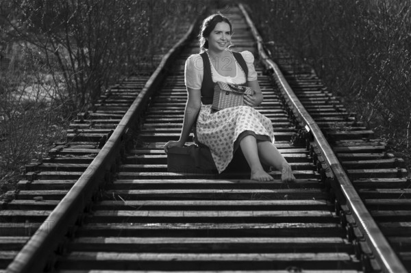 Woman in traditional dirndls sits on railway tracks, relaxed and smiling, black and white picture, Mindelheim, Unterallgäu, Bavaria, Germany, Europe