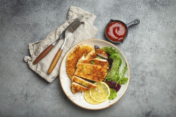 Crispy panko breaded fried chicken fillet with green salad and lemon cut on plate on gray rustic concrete background table with ketchup from above. Japanese style deep fried coated chicken breasts, food photography, food photography
