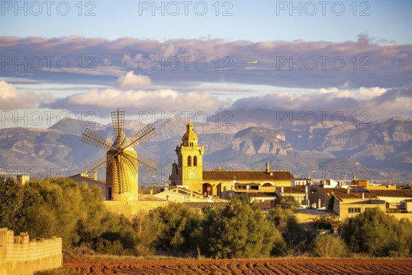 Mountain landscape with village, windmill and church tower under a cloudy sky and flying aeroplane over the mountains, Algaida, Majorca, Spain, Europe